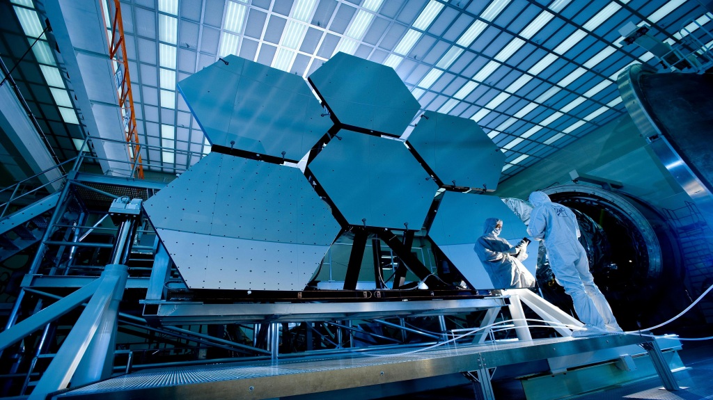 Two scientists in white suits stand near satellite dish-like instruments inside a space station-like facility.