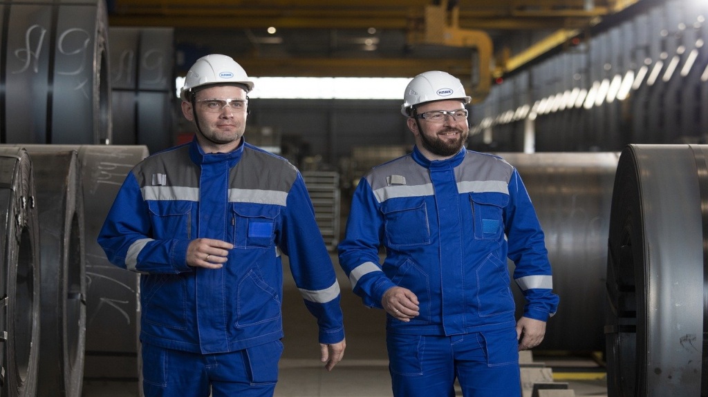 Two middle-aged workers in blue uniforms and helmets smiling in a factory.