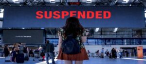 Havalimanında “SUSPENDED” yazılı ekrana bakan yolcu; göçmen vizeleri sürecindeki kısıtlamaları simgeliyor.