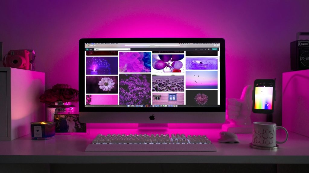 A dimly lit room with purple lights, an open laptop showing images of flowers and raindrops, a phone, a glass of water, and framed photos of a young couple.
