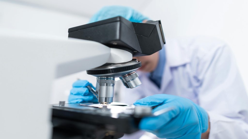 A scientist wearing gloves, a blue head cover, and a white lab coat looks through a microscope.