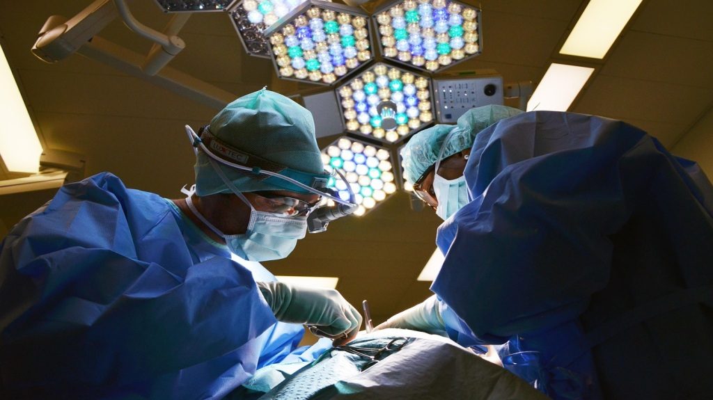 Three doctors in blue scrubs performing surgery under colorful operating lights.