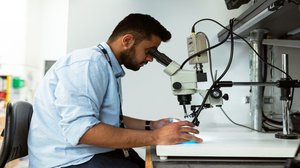 A young man sitting and looking through a microscope in a lab.