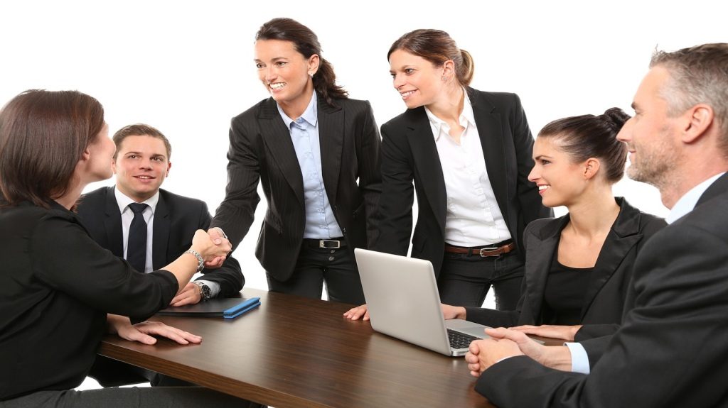 Two women shaking hands while others smile around a conference table.