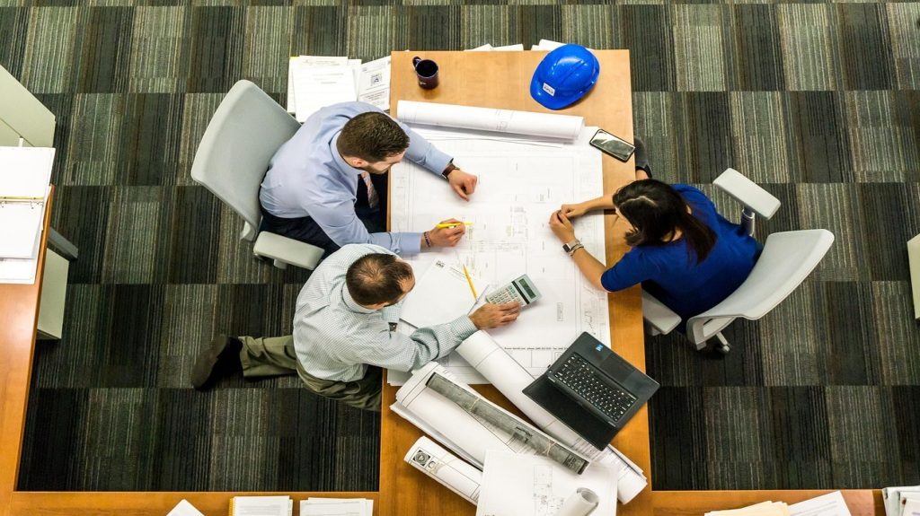 Three people, including one woman, looking at large construction plans on a desk with a helmet, computer, and calculator.