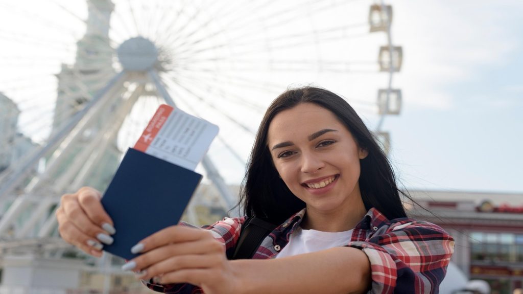 A cheerful young woman showing her passport and boarding pass while smiling at the camera.